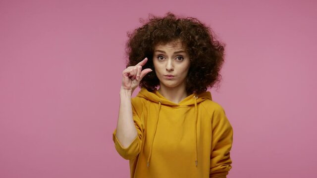 Too small! Girl afro hairstyle in hoodie showing a little bit sign, looking with imploring grimace and disappointed in size, need some more, gesturing minimum. studio shot isolated on pink background