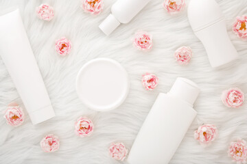 Different white toiletries and pink roses on fluffy fur blanket background. Care about face, hands, legs and body skin. Daily women beauty products. Flat lay. Closeup. Top down view.