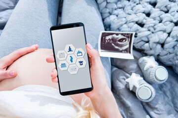 Closeup big female pregnant belly tummy. .Ultrasound picture and little booties are on background. Young woman girl is holding smartphone with interface of maternity application on screen.