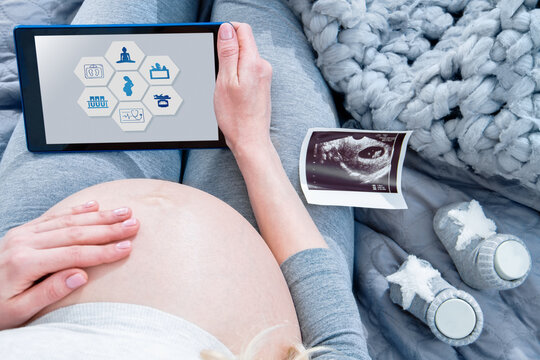 Closeup big female pregnant belly tummy. .Ultrasound picture and little booties are on background. Young woman girl is holding tablet computer with interface of maternity application on screen.