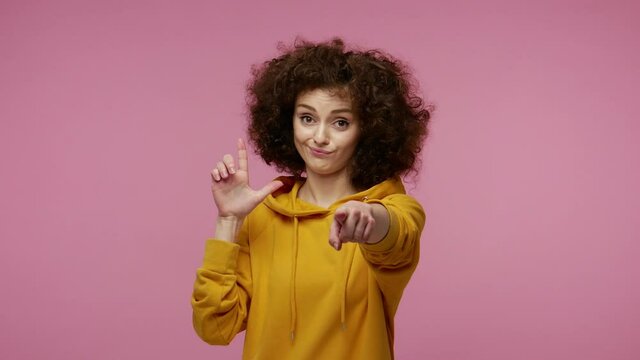 Girl afro hairstyle in hoodie showing loser gesture and pointing finger to camera, expressing disrespect, accusing blaming for failure, life troubles. indoor studio shot isolated on pink background
