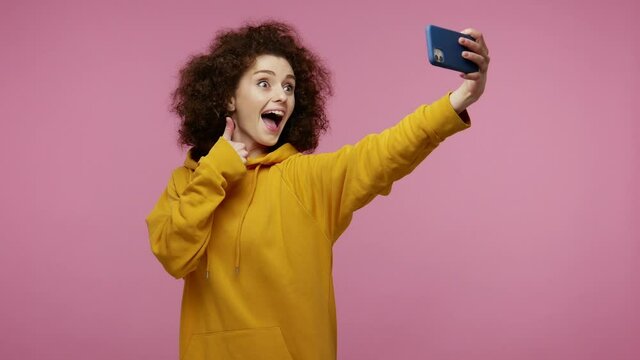 Amazing cheerful stylish girl afro hairstyle in hoodie taking selfie with positive gesture of victory, thumbs up, like, hello and sending air kiss to webcam. studio shot isolated on pink background