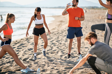 group of young adults doing exercises on sandy beach. beardy guy drinking water from a plastic bottle