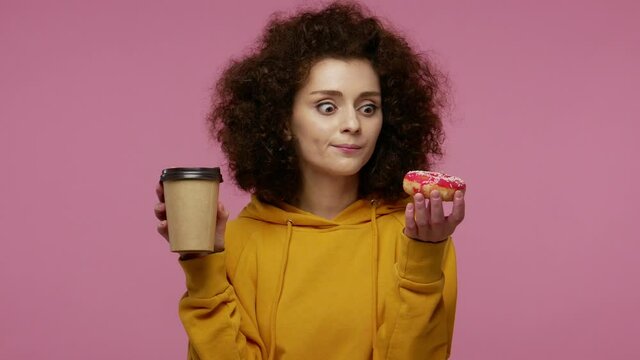 Girl afro hairstyle in hoodie drinking cup of coffee and looking with great desire at donut, craving to bite sweet confectionery, sugar glucose and junk food concept. indoor studio shot isolated