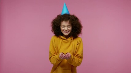 Optimistic girl afro hairstyle in hoodie with funny cone on her head laughing, moving in dance and blowing confetti glitters, celebrating birthday congratulating on anniversary. studio shot isolated