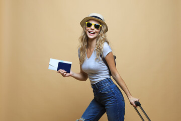 Cheerful blonde woman in sunglasses posing with baggage and holding passport with tickets over beige background.