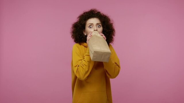 Frightened girl afro hairstyle in hoodie exhaling and inhaling into paper package, using bag with carbon dioxide to improve well-being, overcoming stress fear. studio shot isolated on pink background