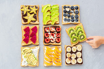 Sandwiches with cream cheese, chocolate, fresh fruits and berries (avocado, blueberries, bananas, kiwi, cherry, apricot, dragon fruit) on a gray concrete background. top view. Flat lay. Healthy diet