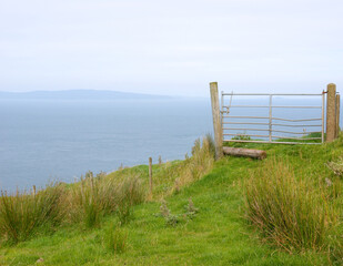 A field gate stands closed on the edge of a cliff overlooking the open sea with blue hills in the distance, illustrating the concept of a road to nowhere, end of the road, no entry, or danger.