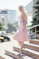 Fototapeta premium A young blonde girl, in a pink dress, descends the stairs of a shopping center on the street