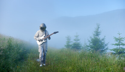 Front view of astronaut in space suit walking on grassy path and playing melody on guitar. Cosmonaut guitarist with musical instrument strolling down foggy meadow with green grass.
