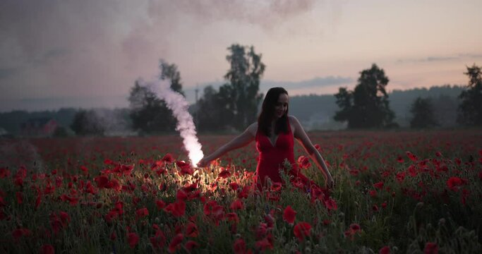 Portrait of pretty brunette girl in a red dress dancing with red burning signal flare in poppy field.
