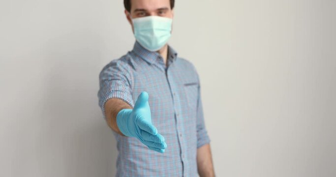 On gray studio background 35s man wears medical face mask and gloves stretch out his hand for handshake. Our new reality due corona virus pandemic outbreak disease. Preventive caution measures concept