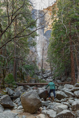 View of the winter Bridalveil Fall, trees, huge river rocks and Tuolumne River in Yosemite Park, California. A man makes his way to the waterfall through rocks and boulders.