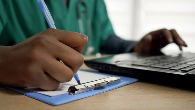 Black doctor works at a computer and makes notes in his office. Close up of doctor hands.