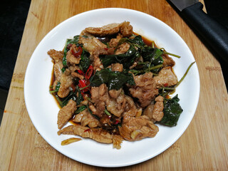 Stir fried beef with holy basil in a white plate, placed on a wooden chopping board and knife. Popular food in Thailand consists of meat, garlic, fresh chilies and basil leaves.