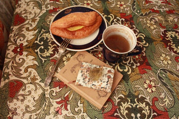 Composition of banana puff on a plate, a blue cup of tea, a fork, two notebooks and a citrine stone on a multi-colored tablecloth.