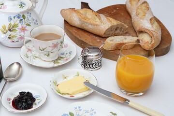 baguette, a cup of tea, orange juice, butter, jam on a white table