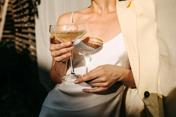 Luxurious woman wearing a white dress, holding a coupe with champagne in sun light. Concept of an...