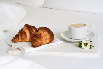 Tasty breakfast with freshly baked croissants and a cup of coffee with lovely flower on the marble tray on the cozy white bed.  Good morning concept. 