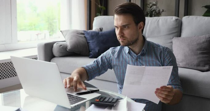 Man working from home holding invoice using calculator and laptop calculates costs, makes payments through e-bank application. Business or household expenses, manage budget incomes or outcomes concept