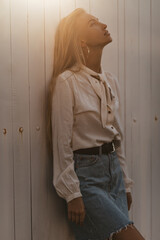 Elegant calm blonde tanned woman in denim stylish skirt and white long-sleeved blouse leans on wooden white wall and looks up.