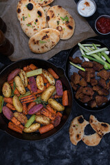 Roasted potatoes and carrots, celery and herbs in cast iron pan on dark table, copy space