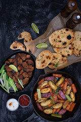 Roasted potatoes and carrots, celery and herbs in cast iron pan on dark table, copy space