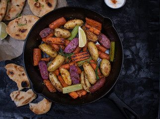 Roasted potatoes and carrots, celery and herbs in cast iron pan on dark table, copy space