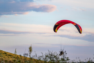 A tandem of people under the red dome of a motor paraglider takes off in the air over a green field...