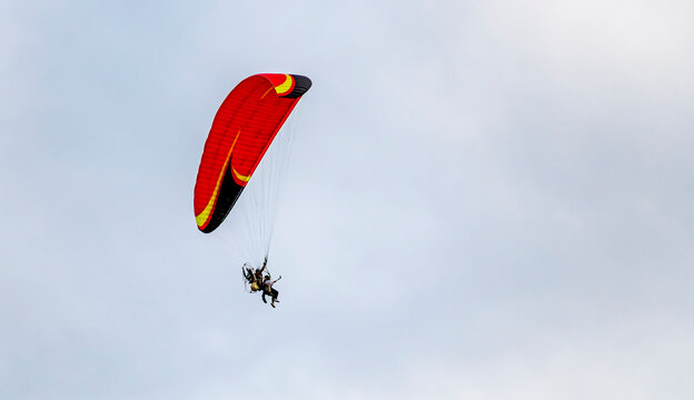 A tandem of people under the red dome of a motor paraglider is flying in the air against a blue sky. Red dome of a motor paraglider against a blue evening summer sky.