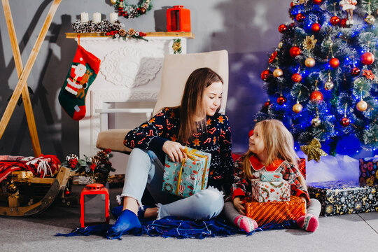 Mom and daughter are sitting under the Christmas tree by the fireplace. New Year's interior. Photo of two sisters under the Christmas tree with gifts.