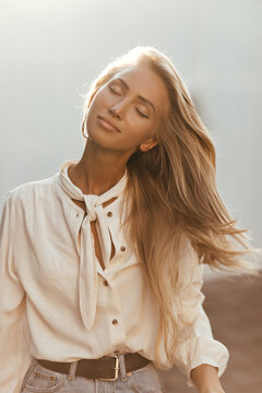 Happy blonde woman in white blouse and denim skirt plays hair. Charming tanned girl poses with closed eyes outside.