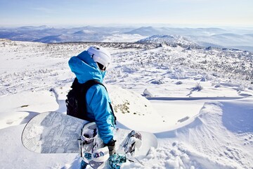 Girl snowboarder is standing on the top of the mountain looking into the distance. Snowboarder getting ready to ride off piste in deep powder snow. Snowboarding in snowy mountains on sunny winter day.
