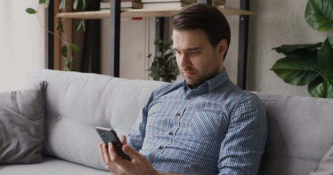Single guy sitting on couch in living room holds cell spend free time in internet chatting with friends, using e-date website online services enjoy distant communication. Web Surfing activity concept