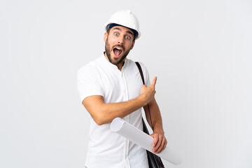 Young architect man with helmet and holding blueprints isolated on white background surprised and pointing side