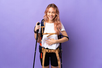 Teenager girl with backpack and trekking poles over isolated purple background smiling a lot