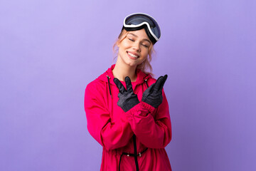 Skier teenager girl with snowboarding glasses over isolated purple background smiling and showing victory sign