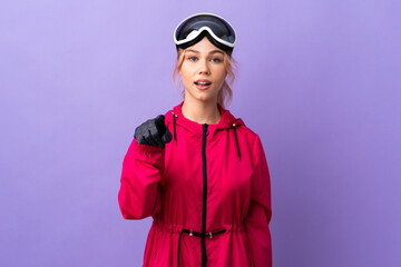 Skier teenager girl with snowboarding glasses over isolated purple background surprised and pointing front