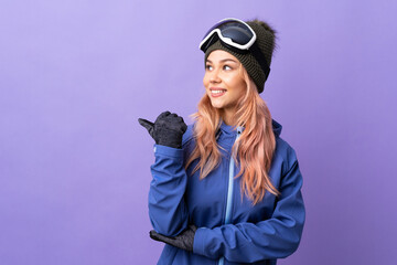 Skier teenager girl with snowboarding glasses over isolated purple background pointing to the side to present a product