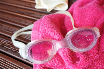 Swim goggles, pink towel and swim cap on wooden deck