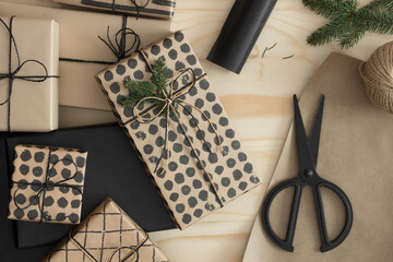 Top view of a kraft and black christmas gifts surrounded with scissors, leaves and twine on a wooden table.
