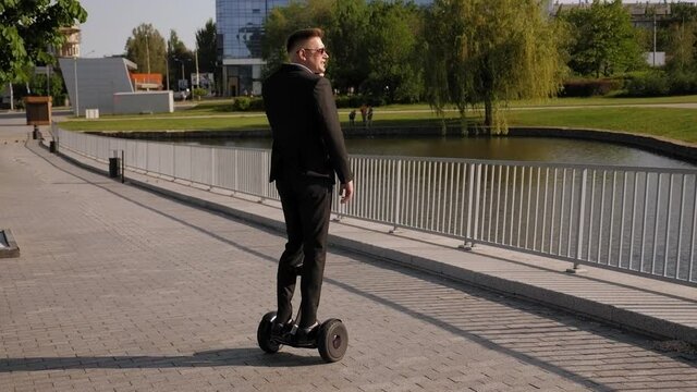 A handsome successful businessman in a black suit and sunglasses rides a gyro scooter in a Park and talks on the phone.