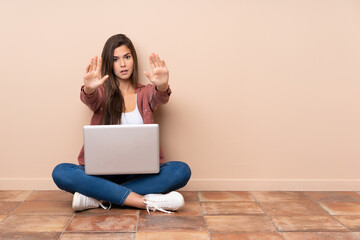 Teenager student girl sitting on the floor with a laptop making stop gesture and disappointed