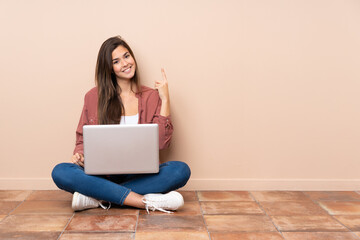Teenager student girl sitting on the floor with a laptop showing and lifting a finger in sign of the best