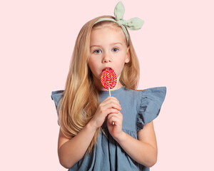 A beautiful European blonde girl with a Lollipop in her hands looks serious on a pink background. Portrait of a beautiful child. Sweets.