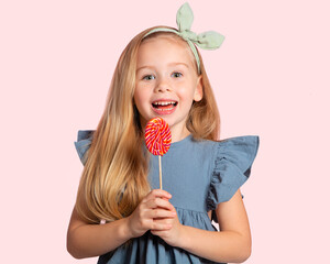 A beautiful European blonde girl with a Lollipop in her hands smiles on a pink background. Portrait of a beautiful child. Happy child. Sweets.