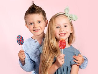 Two children-a beautiful blonde European girl and a boy with blue eyes with lollipops in their hands smile on a pink background. Portrait of beautiful children. Sweets.