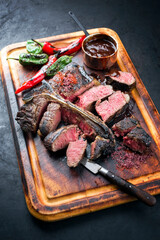 Barbecue dry aged wagyu porterhouse beef steak sliced with large filet piece with chili and hot spice sauce offered as close-up on an old burnt wooden cutting board
