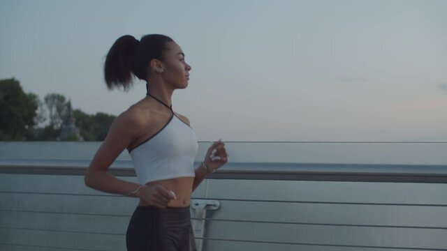 Beautiful motivated african american female jogger with ponytail in sportswear running in the city in early morning. Concentrated sporty fitness woman practicing outdoor workout, jogging at sunrise.